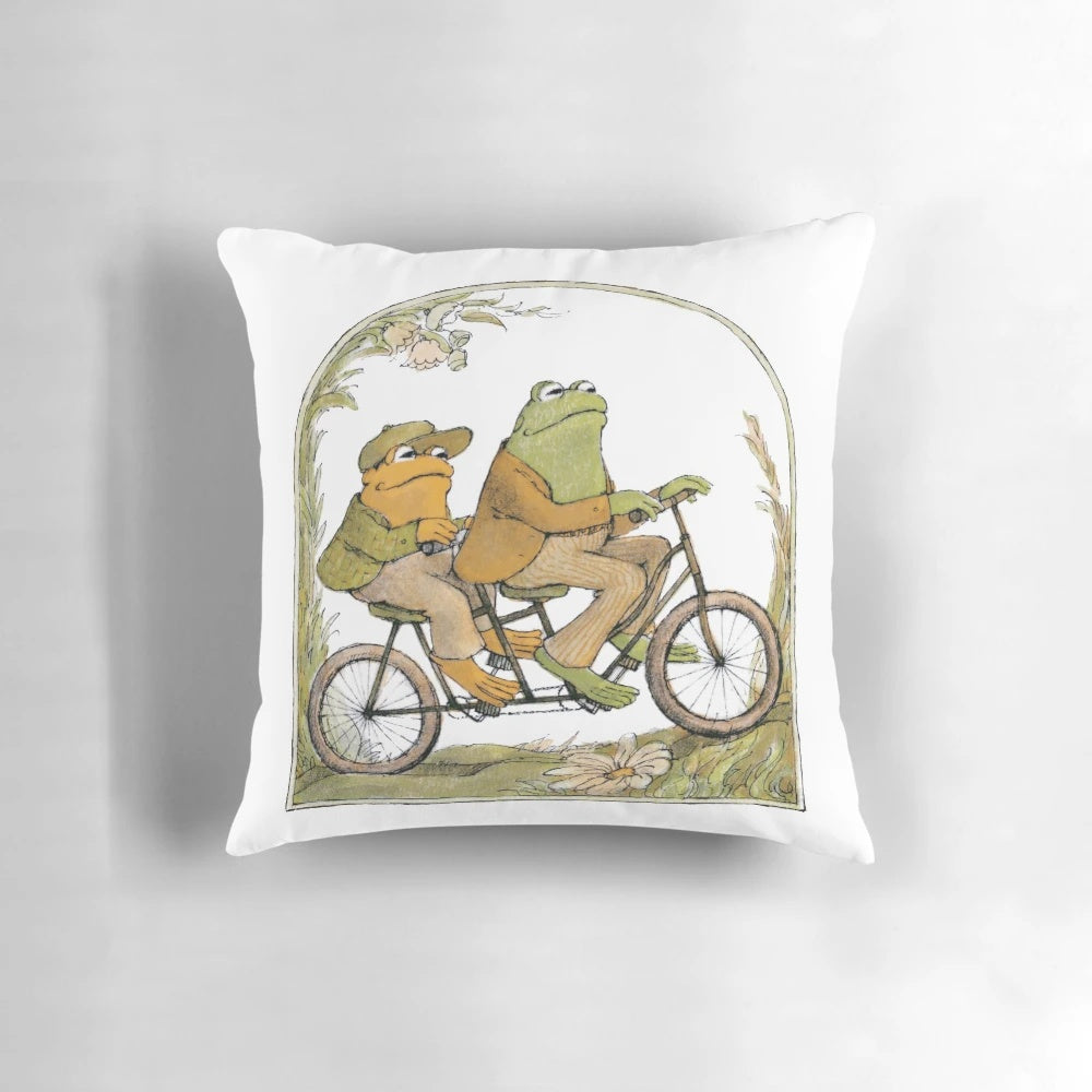 A charming pillow featuring two friends, a frog and a toad, biking together in a whimsical, cottagecore-inspired design.