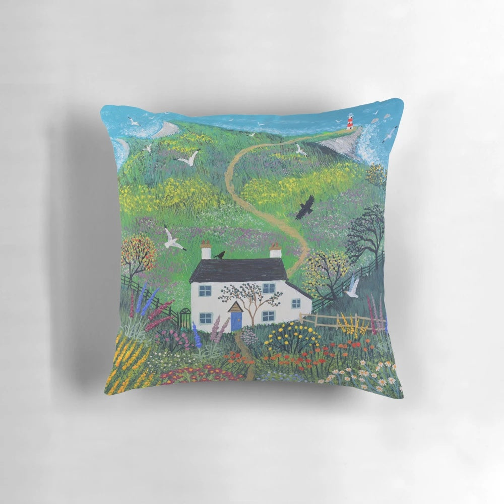 A decorative pillow featuring a serene lighthouse scene against a backdrop of a white cottage, lush gardens, and the sea.