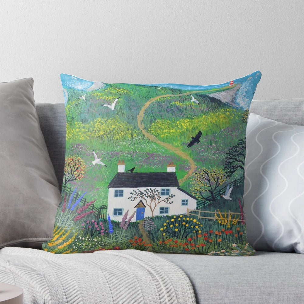 A decorative pillow featuring a serene lighthouse scene against a backdrop of a white cottage, lush gardens, and the sea.