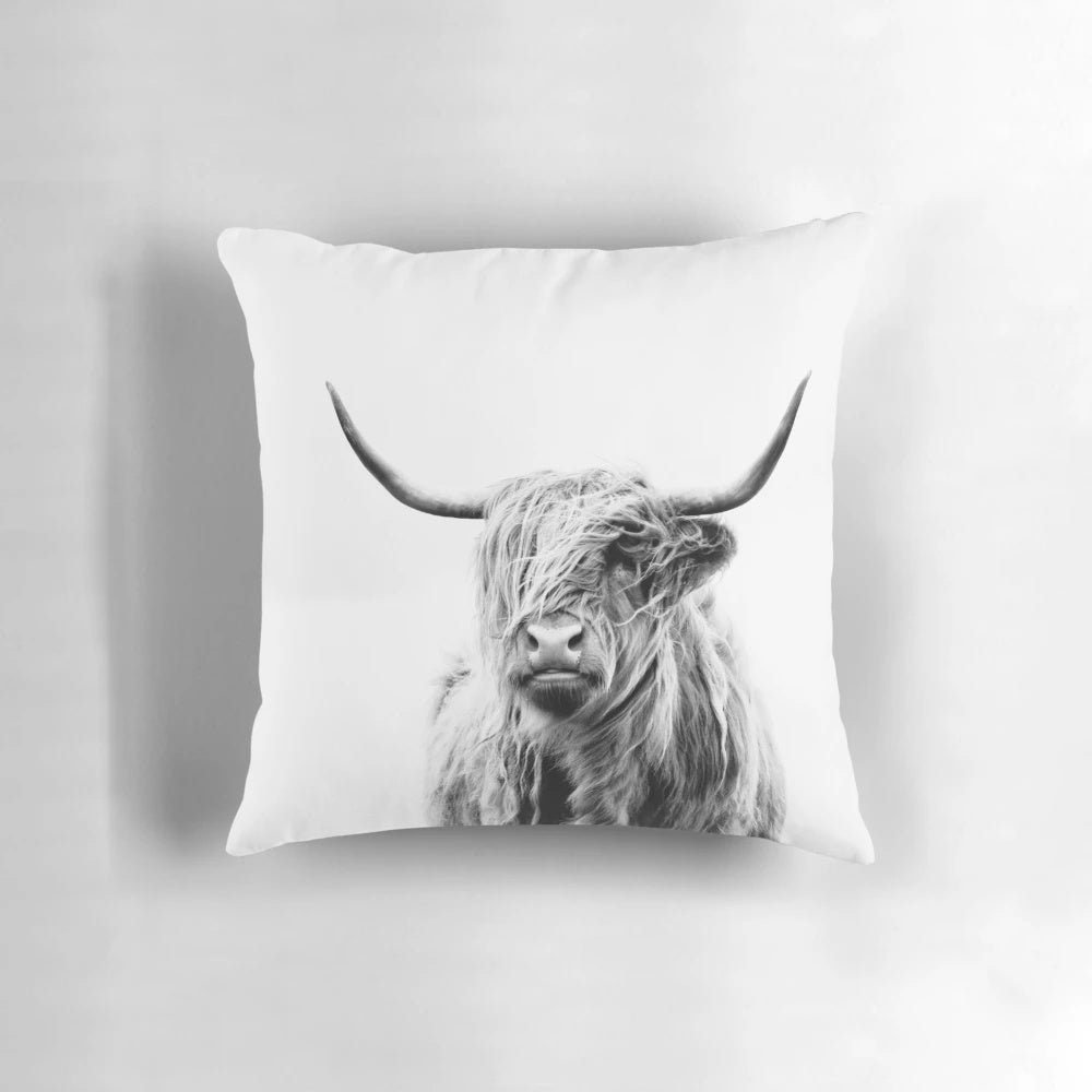 A cozy pillow featuring a portrait of a fluffy, horned highland cow against a warm, rustic backdrop.