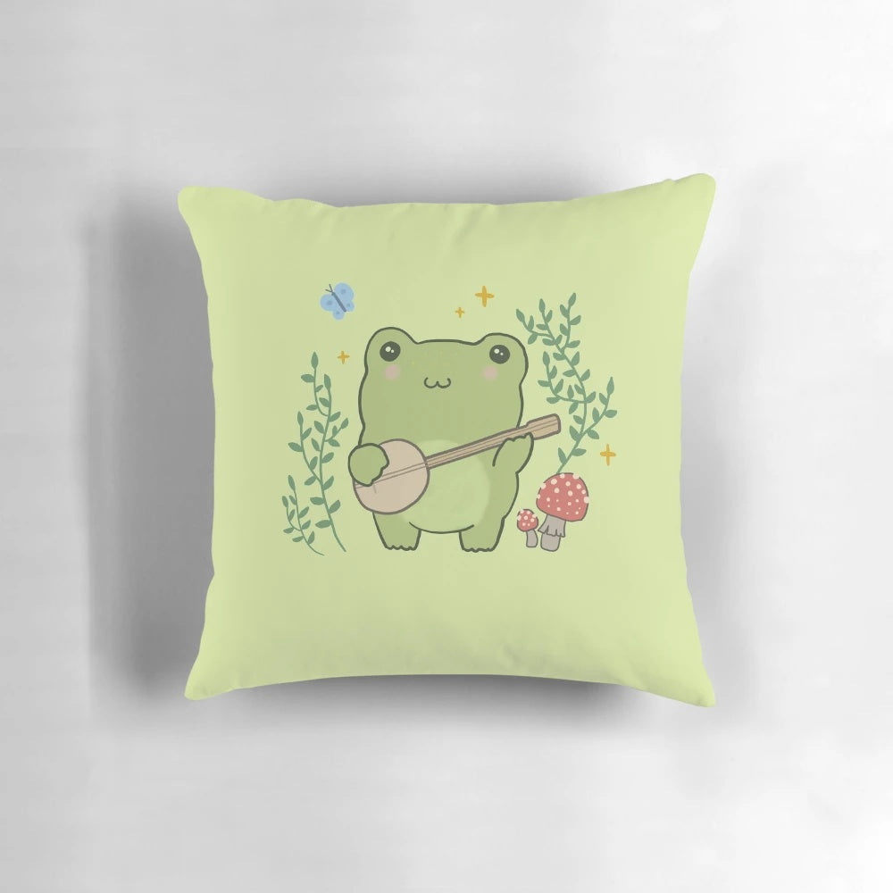 A decorative throw pillow featuring a whimsical, kawaii illustration of a frog playing a banjo, surrounded by mushrooms and a blue butterfly, in a vintage cottagecore aesthetic.