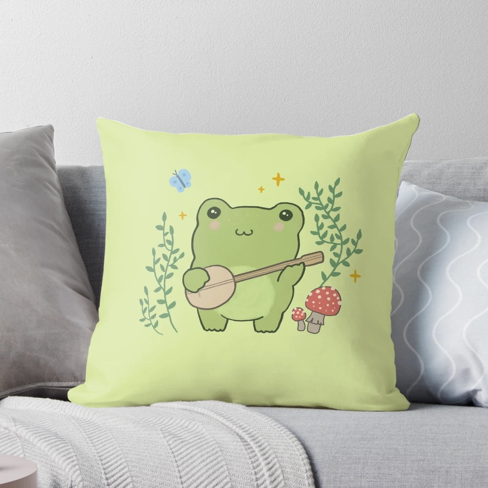A decorative throw pillow featuring a whimsical, kawaii illustration of a frog playing a banjo, surrounded by mushrooms and a blue butterfly, in a vintage cottagecore aesthetic.