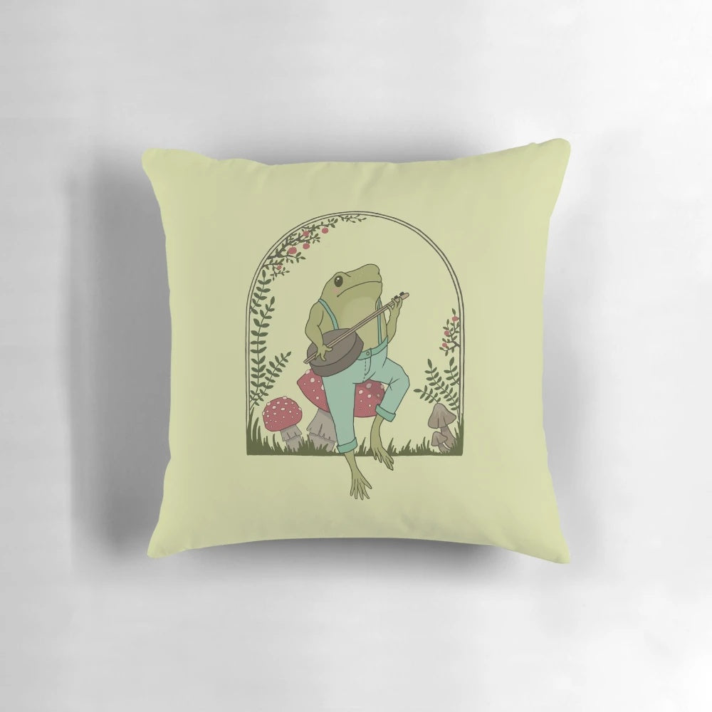 A whimsical cottagecore-style pillow design featuring a frog playing a banjo on a mushroom in a vintage, goblincore-inspired garden setting.