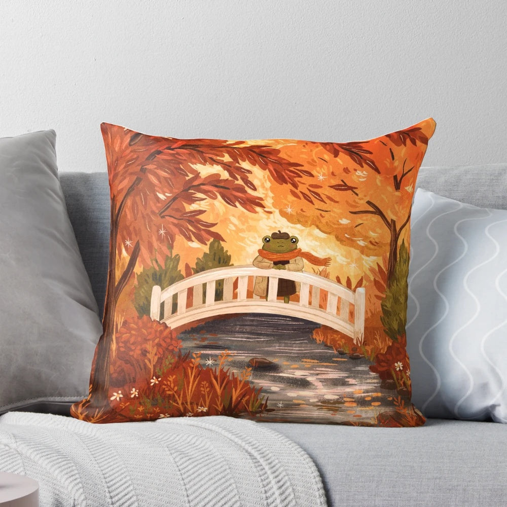 A plush, fall-themed pillow featuring a charming illustration of a frog sitting on a bridge surrounded by vibrant autumn leaves.
