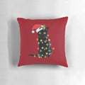 A black cat tangled up in Christmas tree lights, sitting on a festive holiday pillow with a Santa hat.