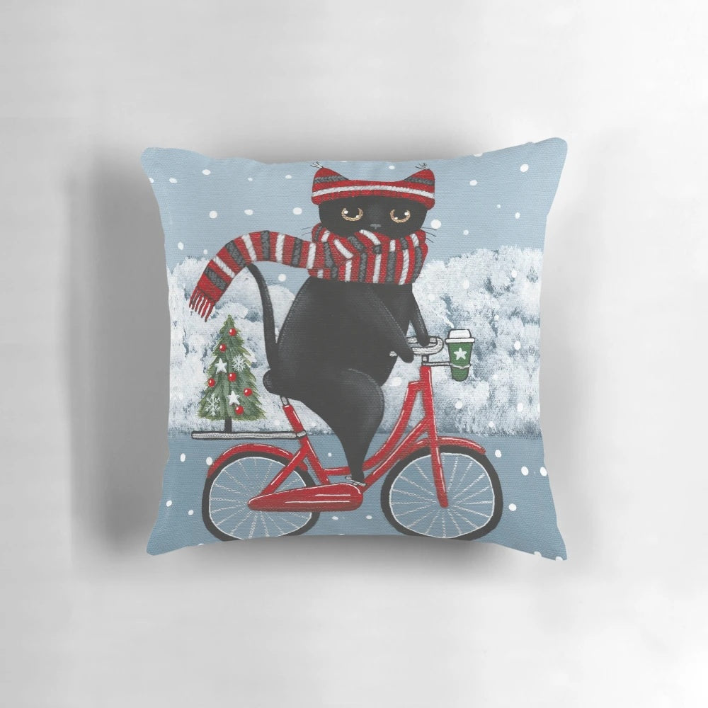 A decorative pillow featuring a black cat riding a bicycle through a winter scene.