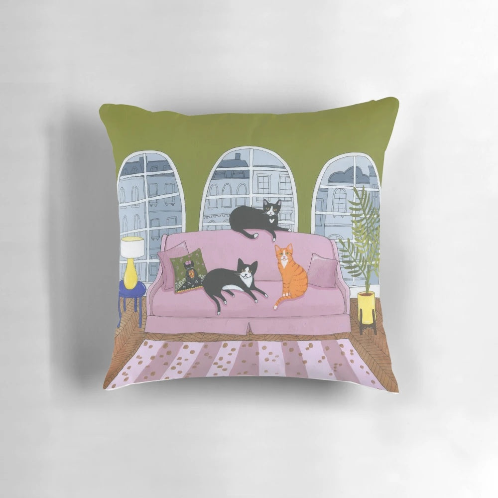 Decorative cat pillow featuring a black, tuxedo, and ginger cat lounging on a pink sofa