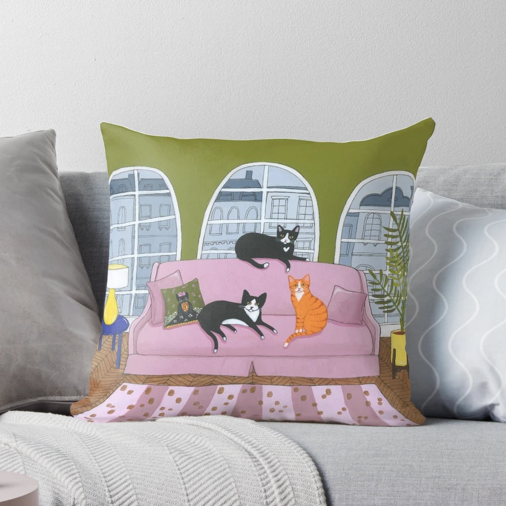Decorative cat pillow featuring a black, tuxedo, and ginger cat lounging on a pink sofa
