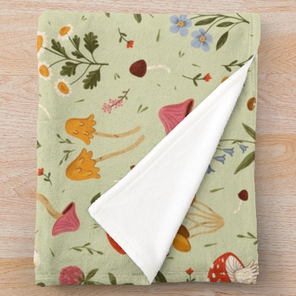 Plush blanket featuring artistic illustration of mushrooms and wildflowers in a natural, woodland setting