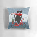 Whimsical tuxedo cat riding a bicycle through a snowy winter scene, printed on a decorative throw pillow.