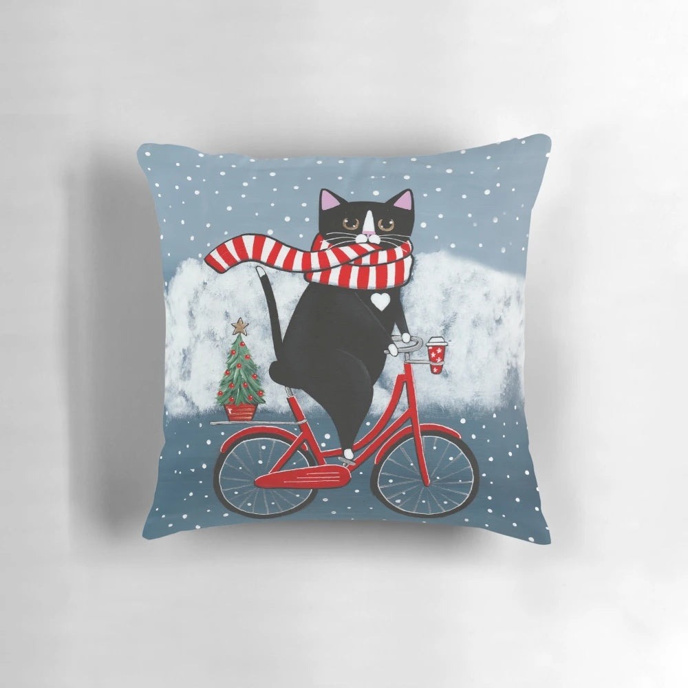 Whimsical tuxedo cat riding a bicycle through a snowy winter scene, printed on a decorative throw pillow.