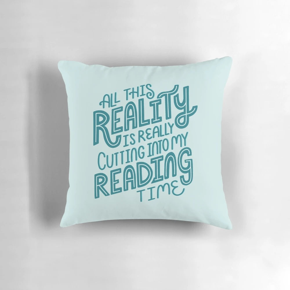 A blue and turquoise throw pillow with the text "Reality Vs. Reading" and a book nerd quote.