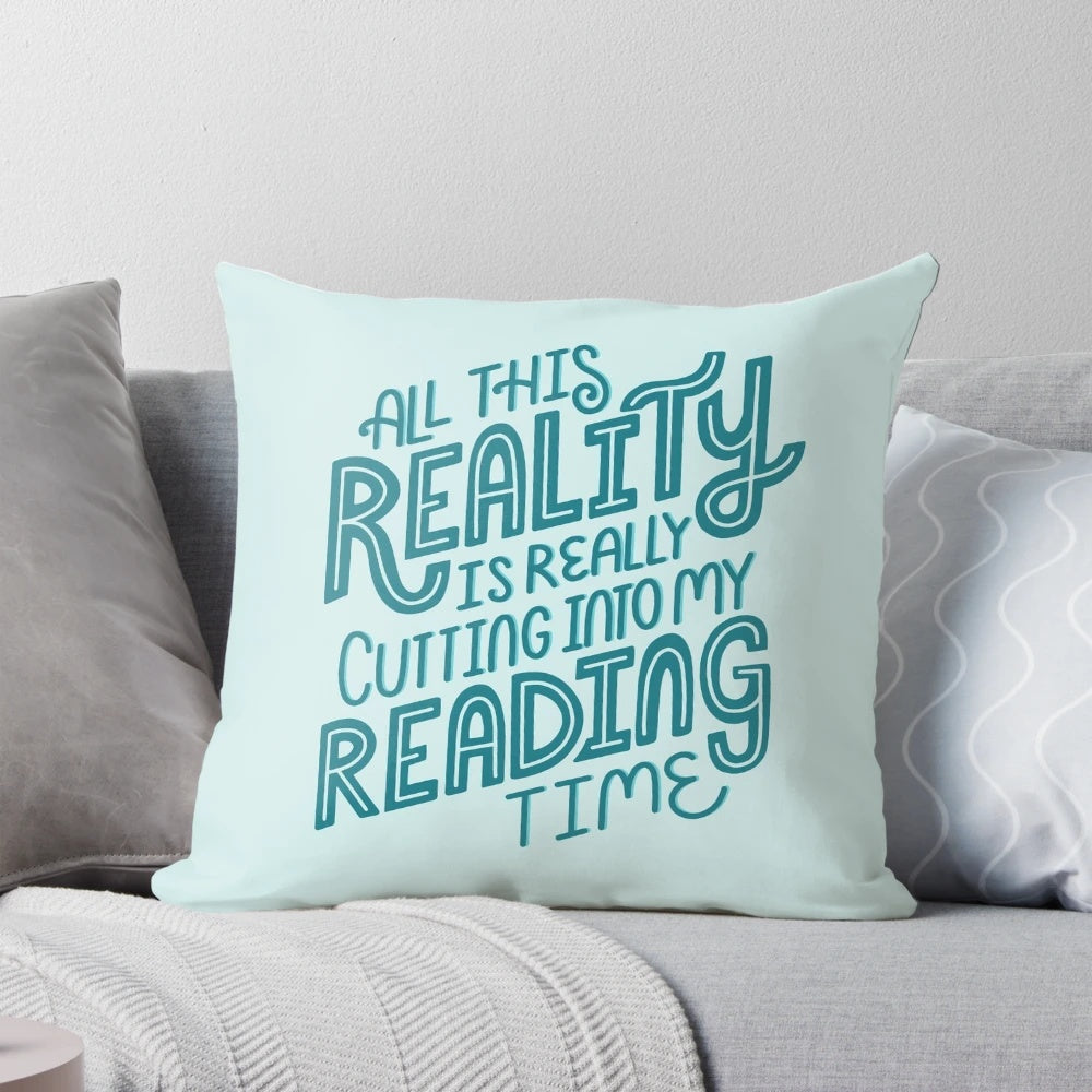 A blue and turquoise throw pillow with the text "Reality Vs. Reading" and a book nerd quote.