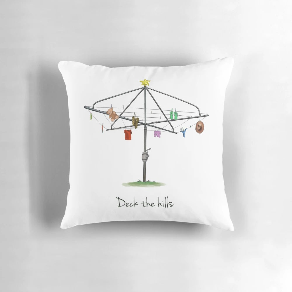 A white square pillow with a whimsical "Deck the Hills" design, featuring Australian imagery like a Hills Hoist clothesline.