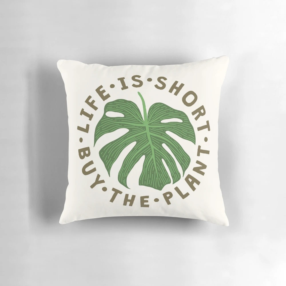 A decorative throw pillow featuring the whimsical message "Life is Short, Buy the Plant" against a lush, tropical plant background.