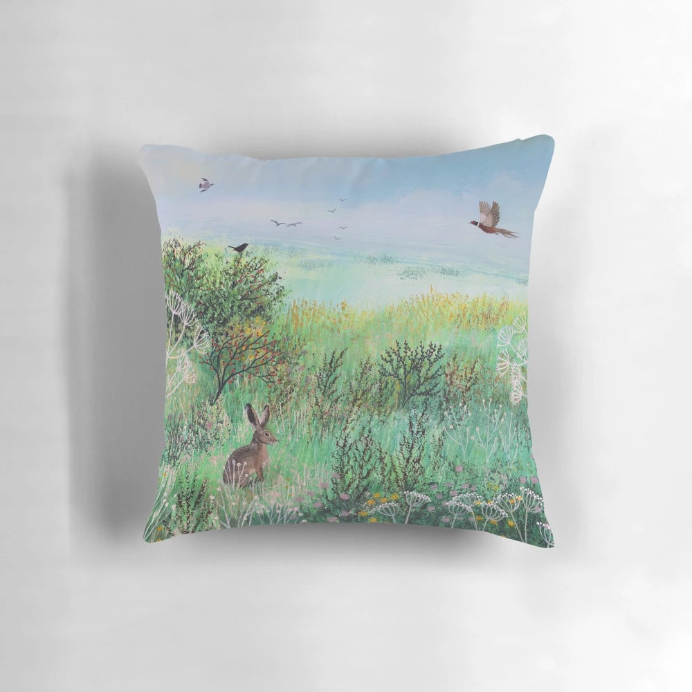 A beautiful pillow featuring a serene meadow landscape with hares and wildlife, designed by artist Jo Grundy.