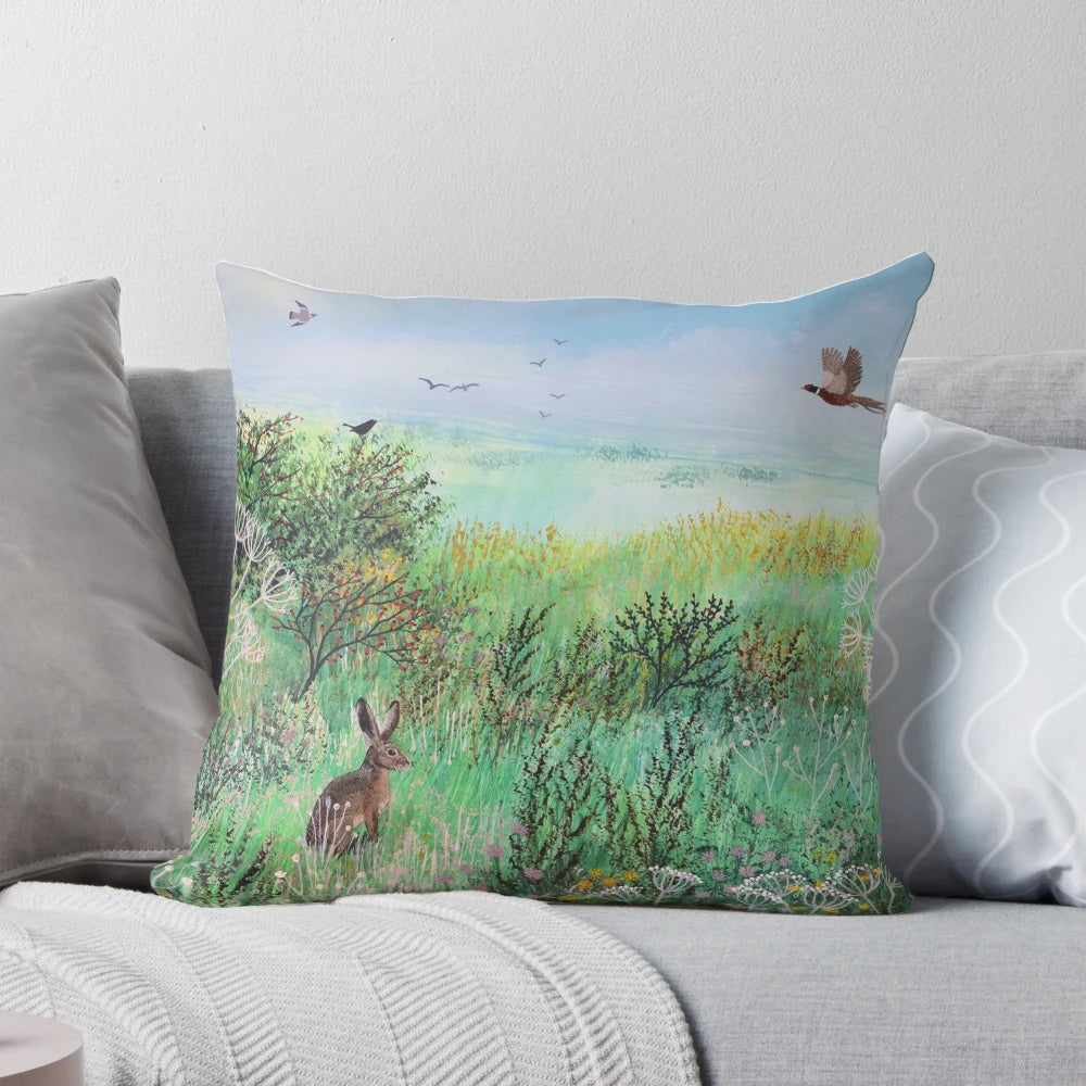 A beautiful pillow featuring a serene meadow landscape with hares and wildlife, designed by artist Jo Grundy.
