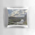 Sunset over a river with trees, guard rails, and rocks on the shore, printed on a square throw pillow