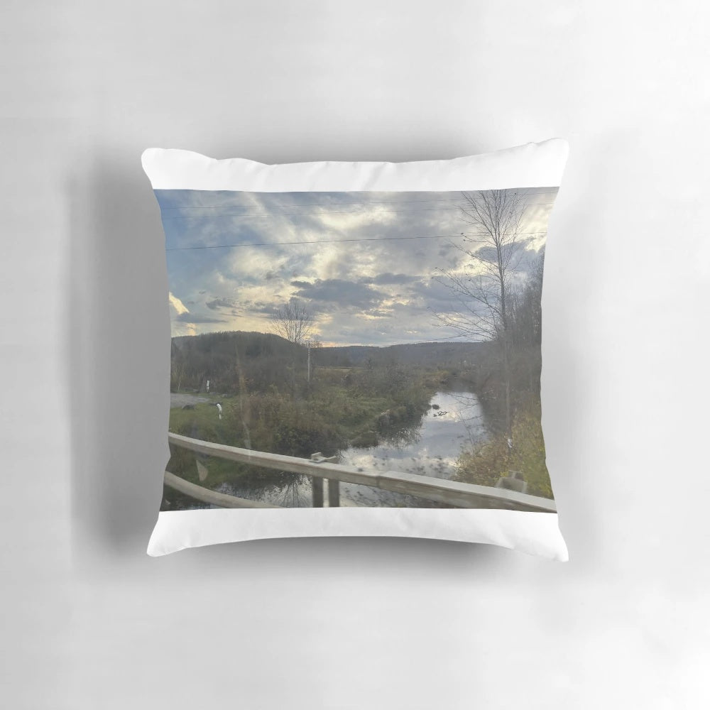 Sunset over a river with trees, guard rails, and rocks on the shore, printed on a square throw pillow