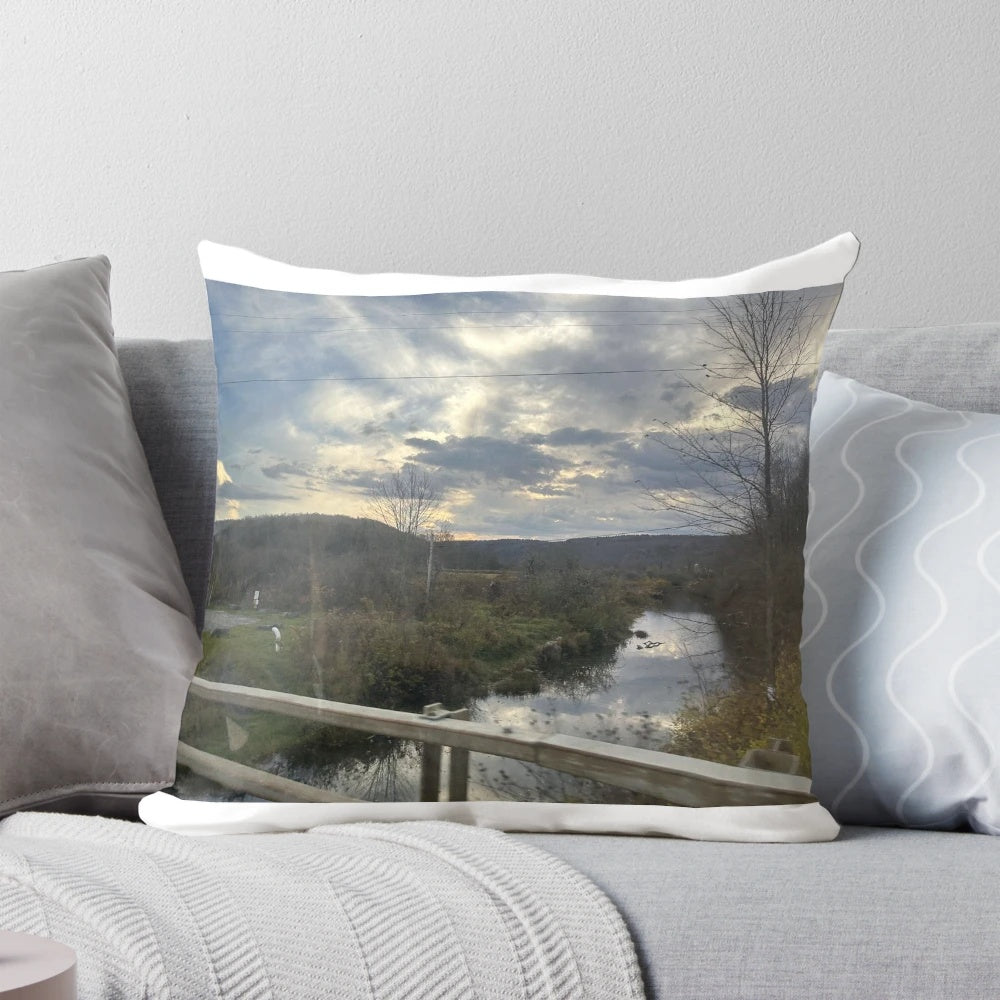 Sunset over a river with trees, guard rails, and rocks on the shore, printed on a square throw pillow