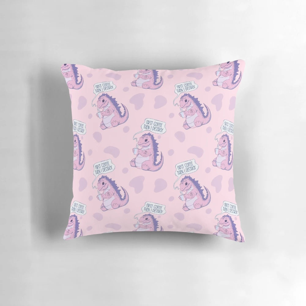 A pastel pink pillow with a playful dinosaur-like monster character and the text "First coffee then I destroy" printed on it.