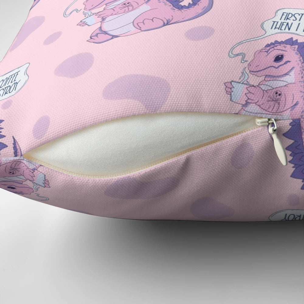 A pastel pink pillow with a playful dinosaur-like monster character and the text "First coffee then I destroy" printed on it.