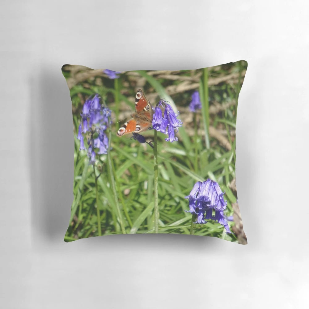 A decorative pillow featuring a vibrant peacock butterfly perched on a bed of blooming bluebells in a lush, wild woodland scene.