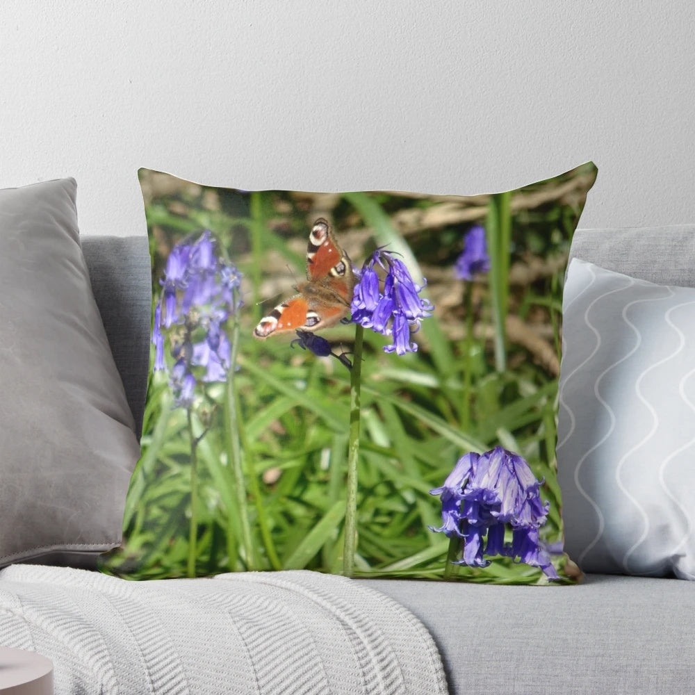 A decorative pillow featuring a vibrant peacock butterfly perched on a bed of blooming bluebells in a lush, wild woodland scene.